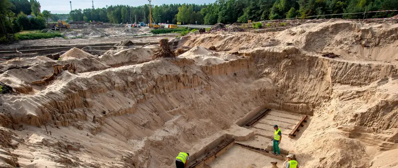 Wykopaliska archeologiczne przed pozwoleniem na budowę odsłaniają drewniane konstrukcje w piasku. Archeolodzy w kamizelkach pracują na miejscu.