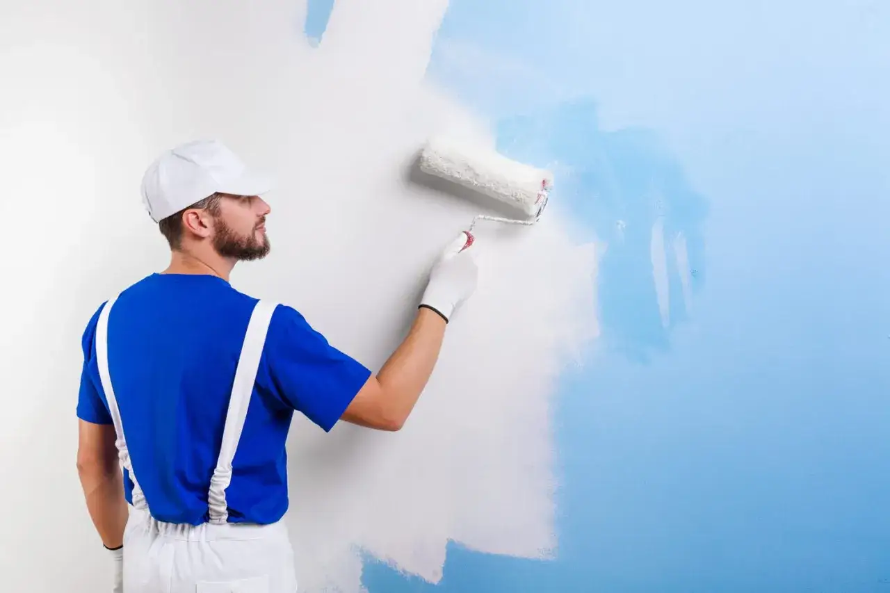 Un peintre en bleu repeint un mur blanc en bleu clair avec un rouleau.