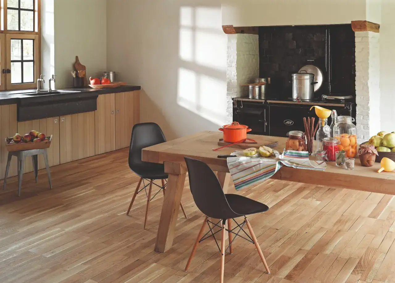 Cocina r&uacute;stica con suelo de madera natural, mostrando diferentes tipos de parquet. Mesa de madera con sillas negras, cocina de le&ntilde;a y encimera con conservas.