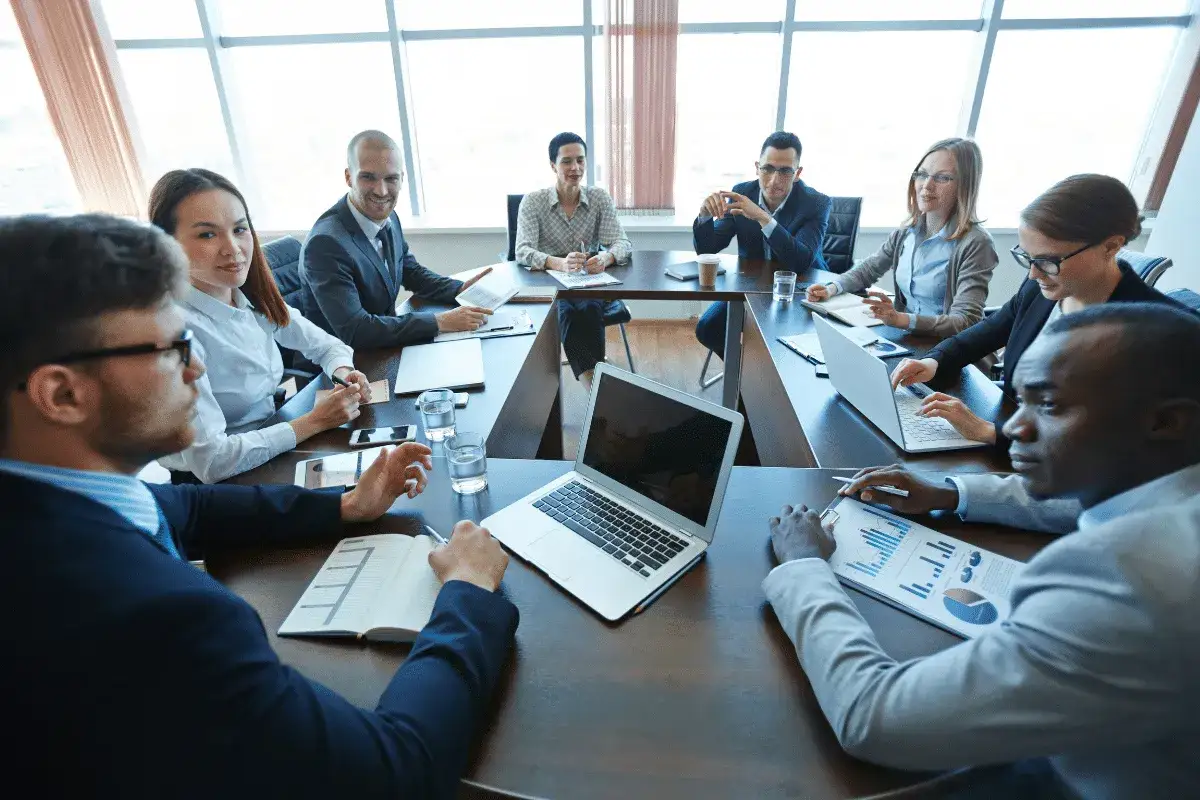 Diverse team in a boardroom, discussing board member requirements. Laptops, papers, and water glasses on the table.