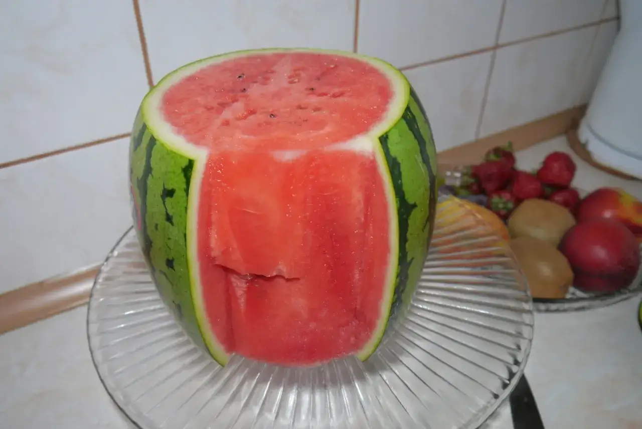 Oryginalny tort z arbuza, z wyciętym środkiem, prezentuje się soczyście. Obok miska z truskawkami i kiwi.