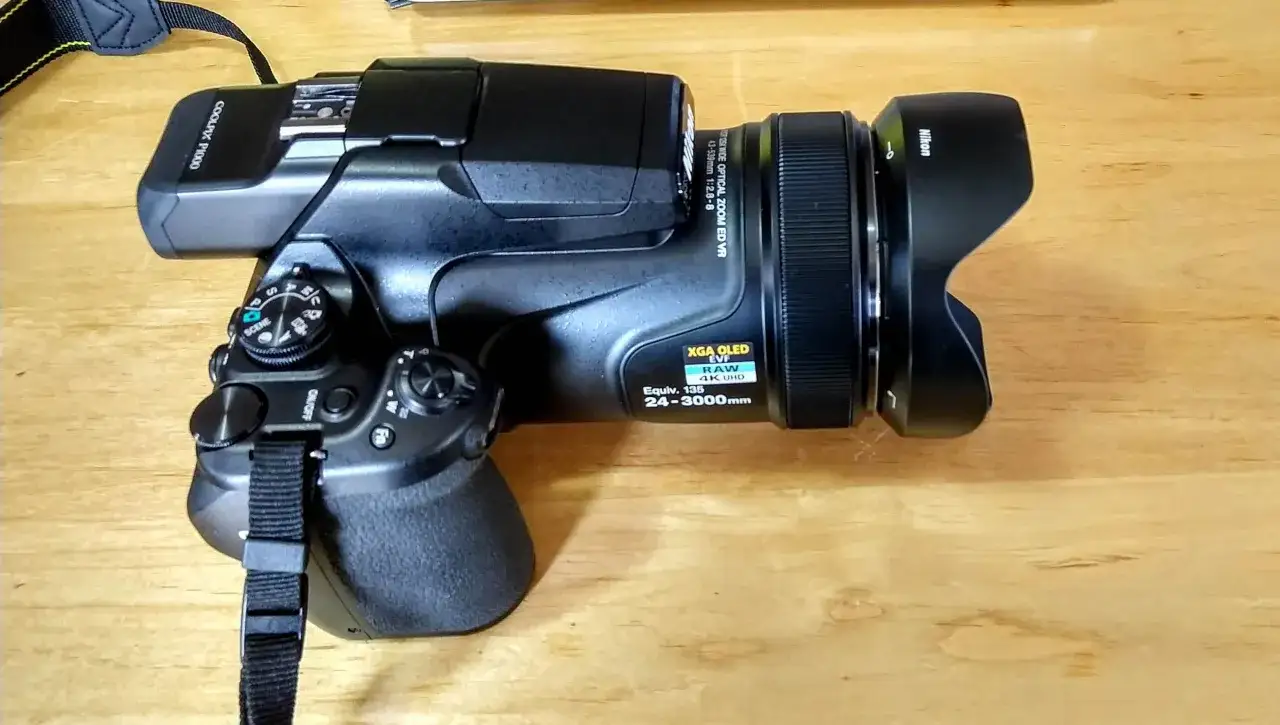 Aparat Nikon P1000 z obiektywem i osłoną przeciwsłoneczną. Widoczny pasek na szyję i pokrętło wyboru trybów.
