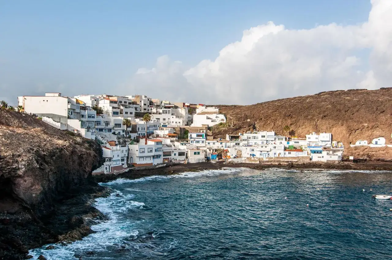 Pueblo blanco de casas apiñadas en la costa rocosa de **Tufia**, con el mar azul y olas rompiendo.