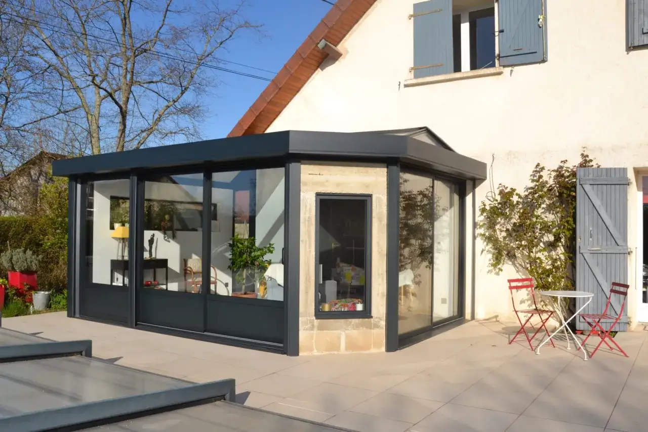 Une v&eacute;randa moderne avec de grandes baies vitr&eacute;es, un petit salon de jardin et une maison en arri&egrave;re-plan. L'assurance veranda est essentielle pour prot&eacute;ger cet investissement.