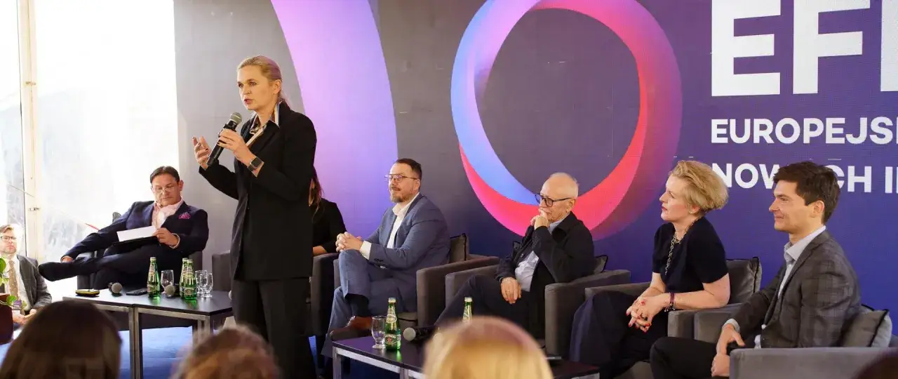 Minister edukacji przemawia na panelu dyskusyjnym. Obok niej siedzą inni uczestnicy, w tym mężczyzna w okularach i kobieta z blond włosami.