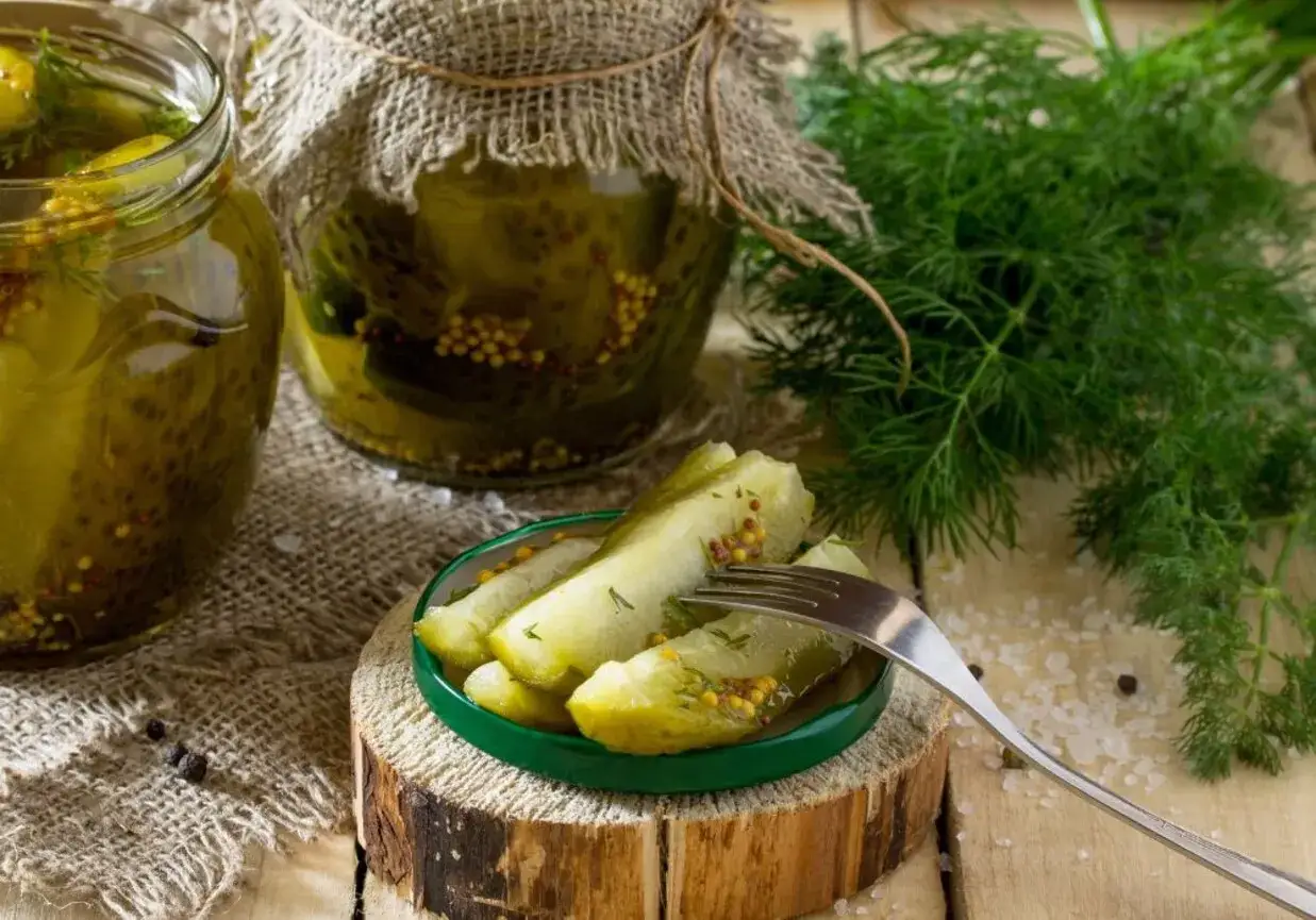 Słoiki z ogórkami w musztardzie, świeży koperek i widelec z ukiszonymi kawałkami. Domowe przetwory gotowe do spożycia.