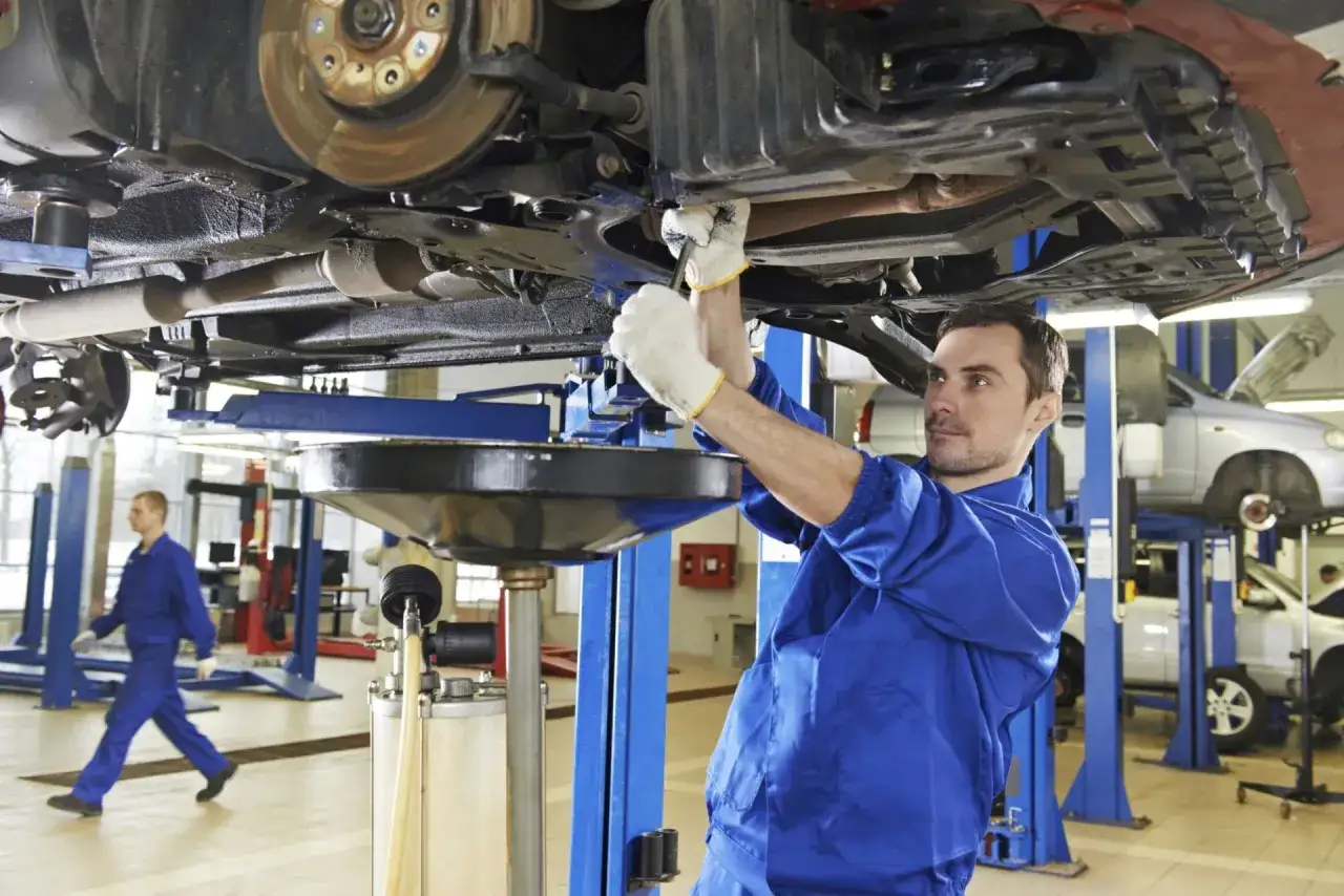 Mechanik samochodowy w niebieskim kombinezonie pracuje pod podniesionym autem. W tle inny pracownik.