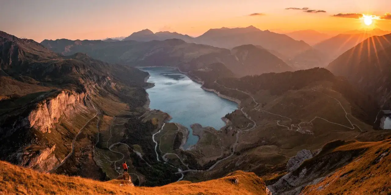 Un randonneur admire le coucher de soleil sur le lac et les montagnes du massif du Beaufortain. Les rayons dorés illuminent les crêtes.