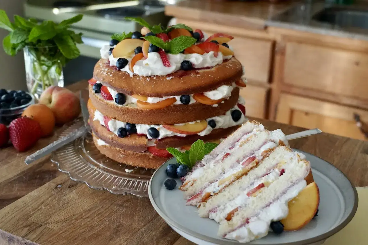Eine üppige Sommer Torte mit frischen Beeren und Pfirsichen, dekoriert mit Minze. Ein Stück ist bereits auf einem Teller angerichtet.