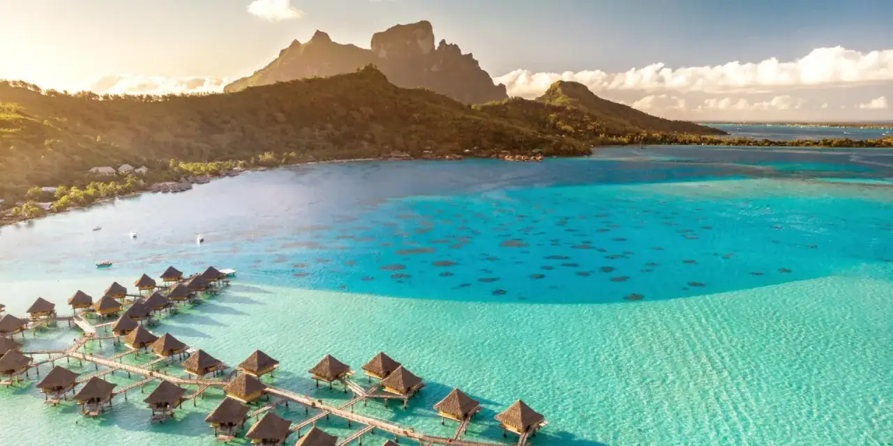 Bungalows sur pilotis dans un lagon turquoise, avec des montagnes verdoyantes en arrière-plan. Un rêve de polynésie française.
