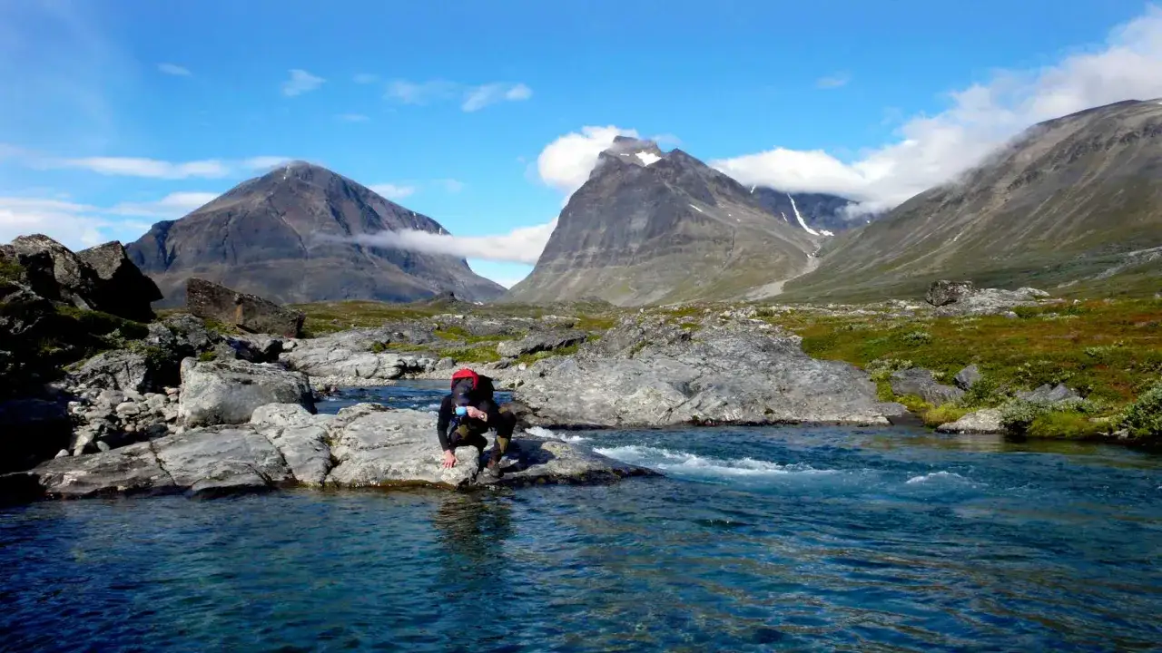 Vandrare vilar vid en forsande fjällbäck under en Kebnekaise vandring. Majestätiska berg reser sig i bakgrunden.