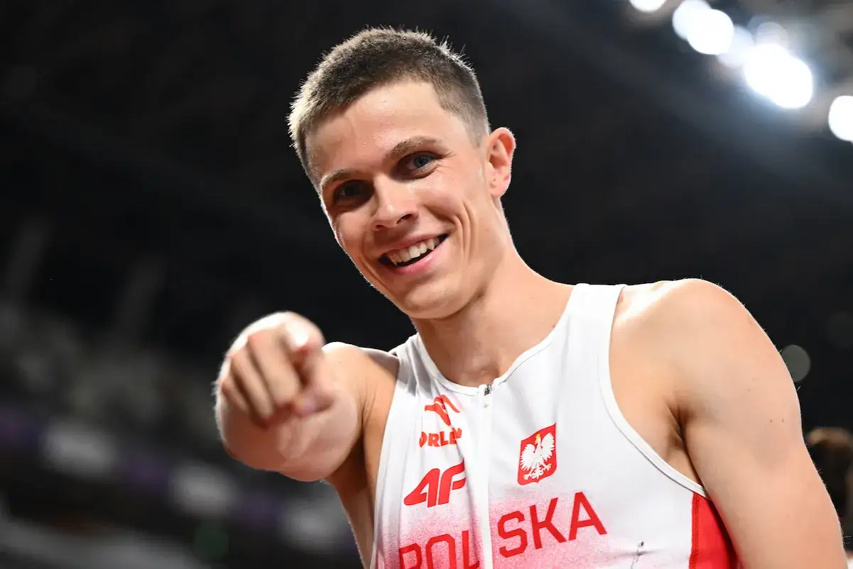 Uśmiechnięty lekkoatleta Maciej Wyderka w biało-czerwonym stroju z orłem, wskazuje palcem w stronę widza.