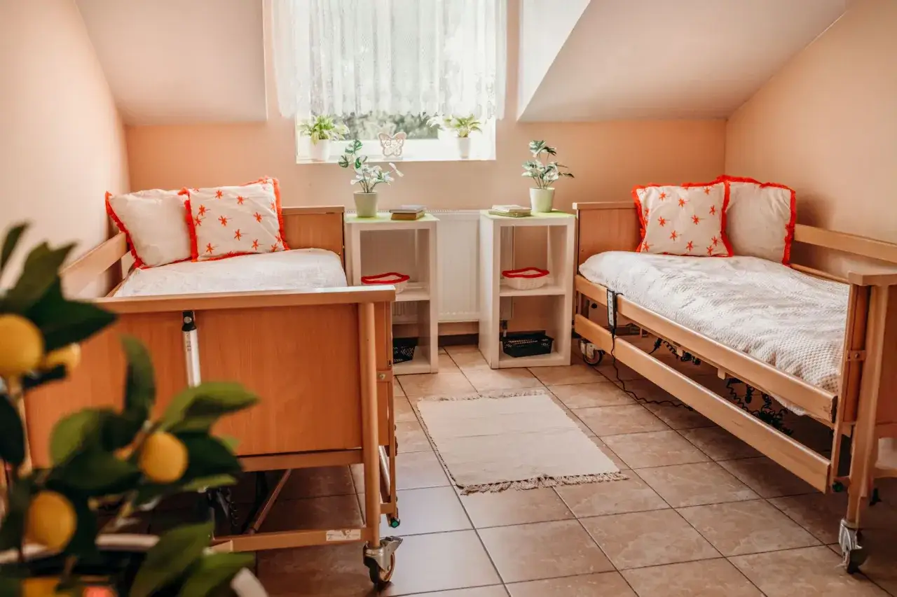 Przytulne pokoje w domu seniora Anna. Dwa łóżka z ozdobnymi poduszkami, stoliki z roślinami i książkami.