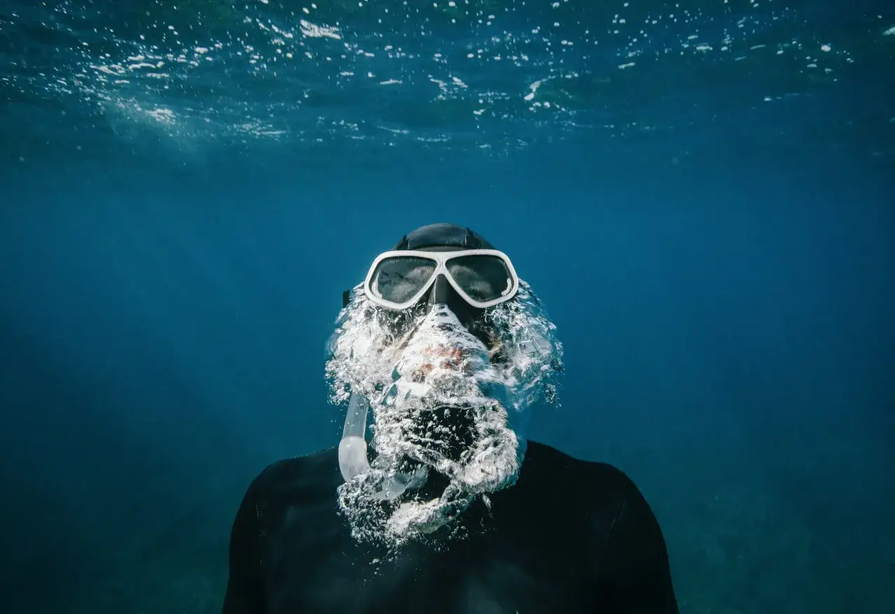 Osoba w masce i czepku zanurza się w wodzie, wydychając bąbelki. To właśnie freediving – nurkowanie na wstrzymanym oddechu.