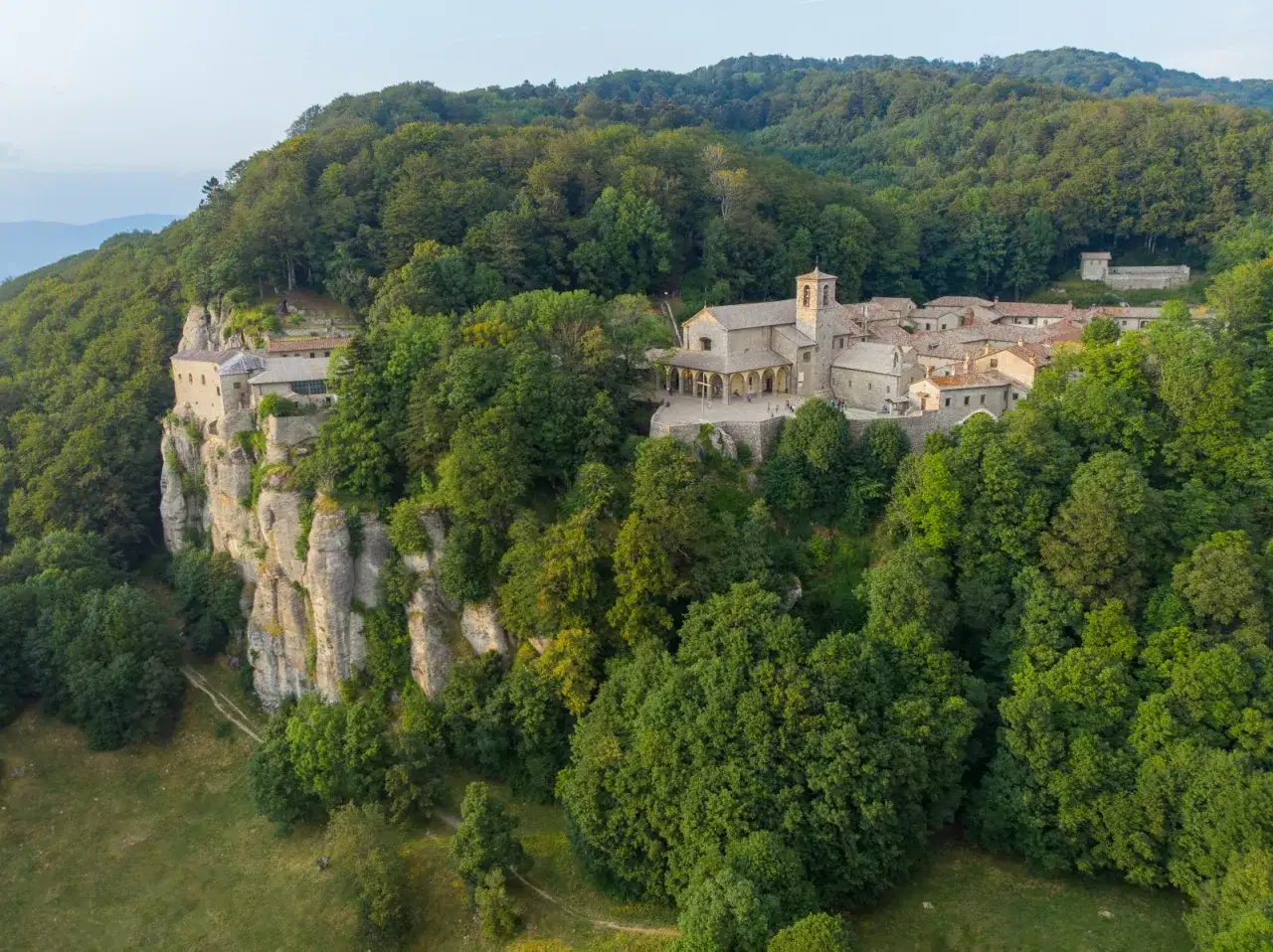 A monastery complex clings to a cliff face in La Verna, Italy, surrounded by lush green forests.