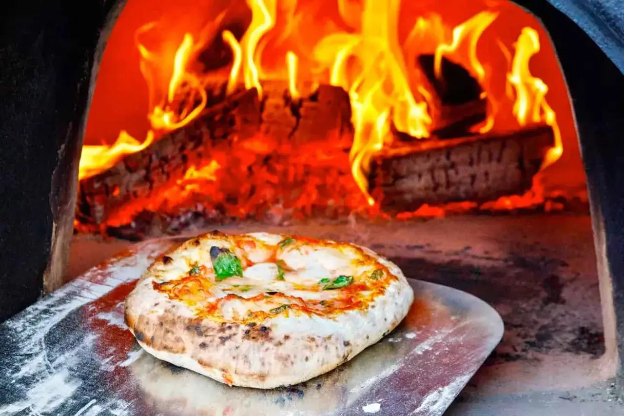 Eine frisch gebackene neapolitanische Pizza mit Mozzarella und Basilikum auf einem Pizzaschieber vor lodernden Flammen im Holzofen.