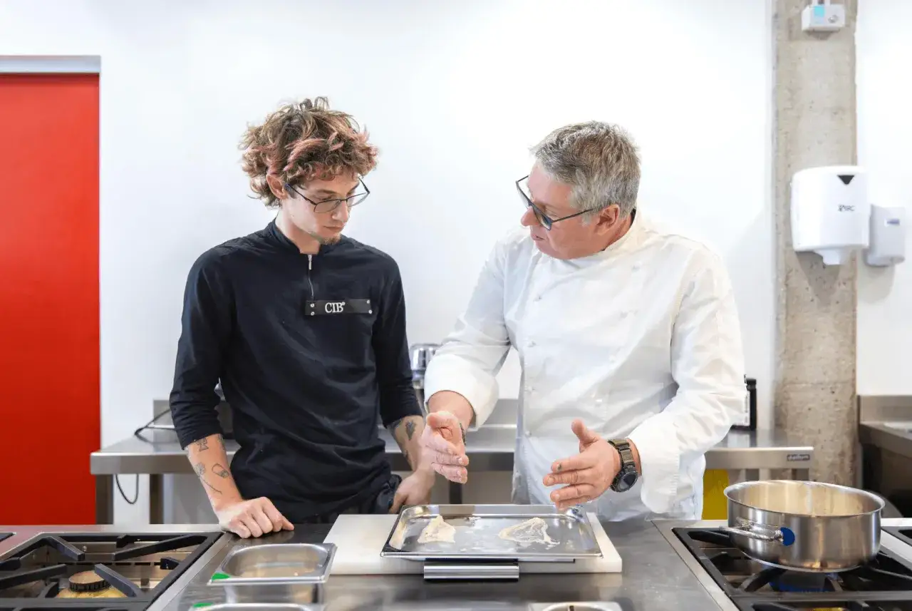 Un chef experimentado explica una técnica a un joven aprendiz. El **convenio jefe de cocina** se refleja en la transmisión de conocimientos.