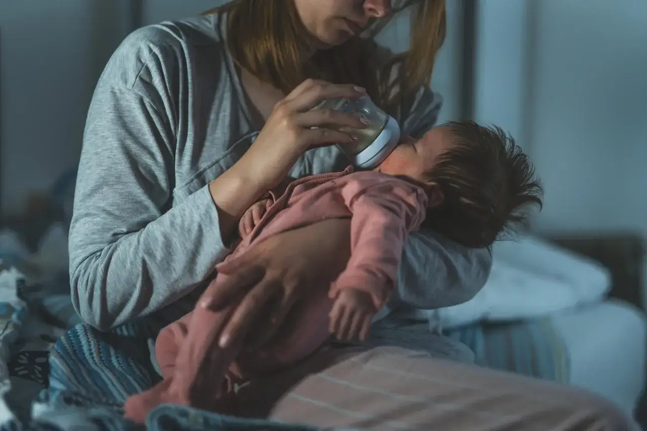 Nocne karmienie uspokaja histeryczny płacz dziecka. Matka tuli niemowlę, podając mu butelkę z mlekiem w półmroku sypialni.