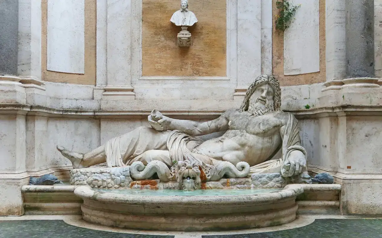 A reclining marble statue of a river god, part of the Capitoline Museums, rests in a fountain with water spouting from a lion's head.