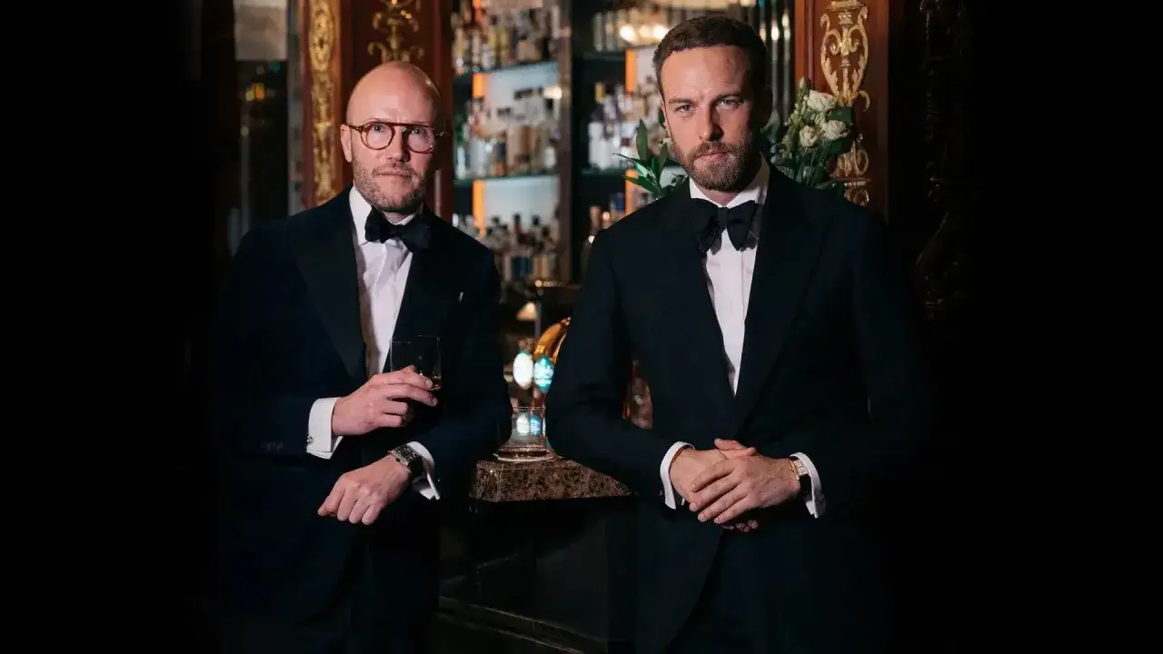 Two men in tuxedos, embodying holiday attire, stand at a bar. One holds a drink, the other rests his hands.