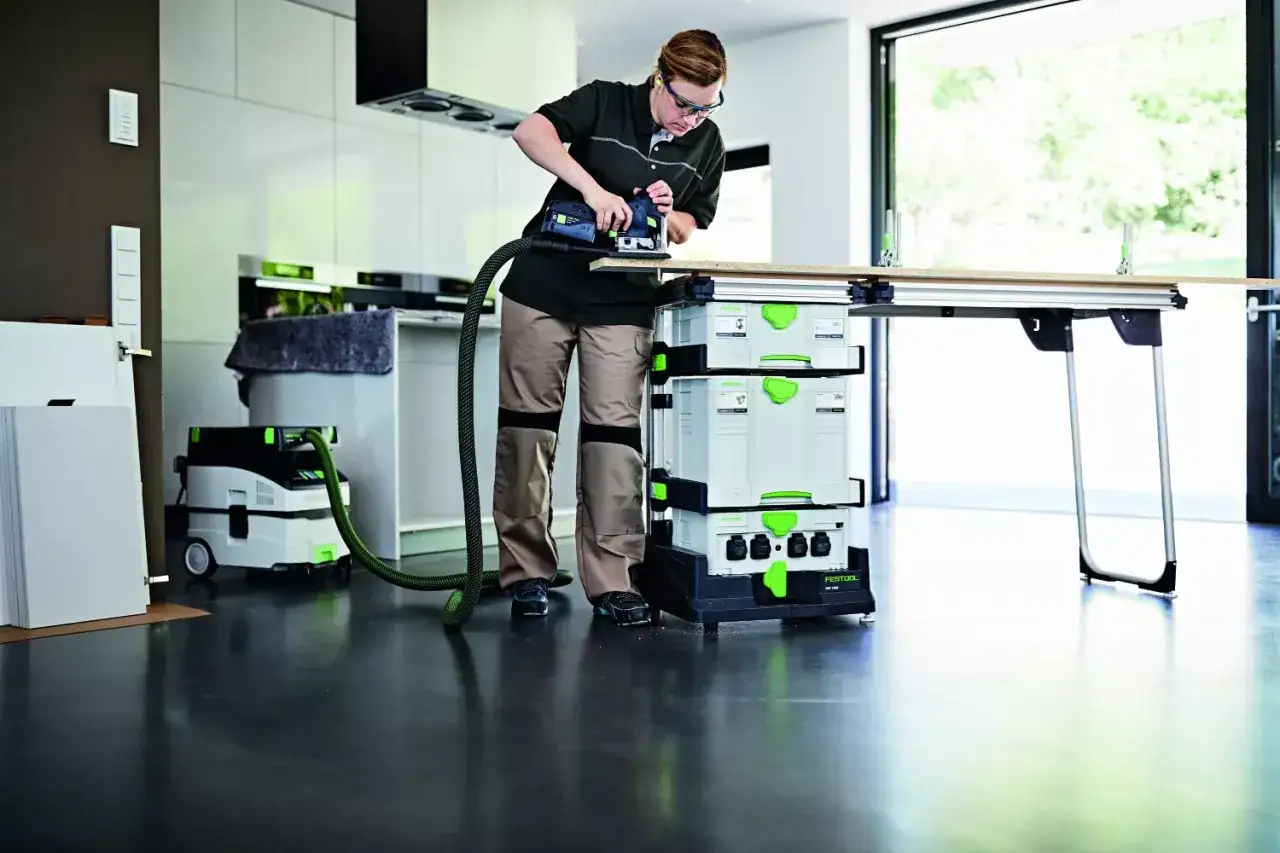 Une femme utilise un ponceuse sur un &eacute;tabli, avec un aspirateur et des bo&icirc;tes de rangement Festool. Son atelier mobile est pr&ecirc;t pour le travail.