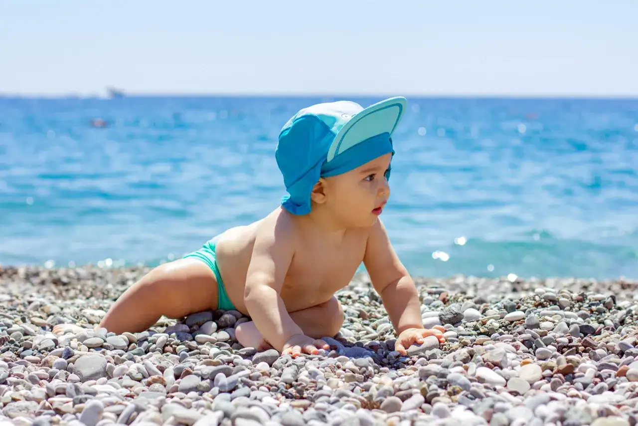 Un neonato con cappellino blu esplora la spiaggia di ciottoli, con il mare azzurro sullo sfondo.