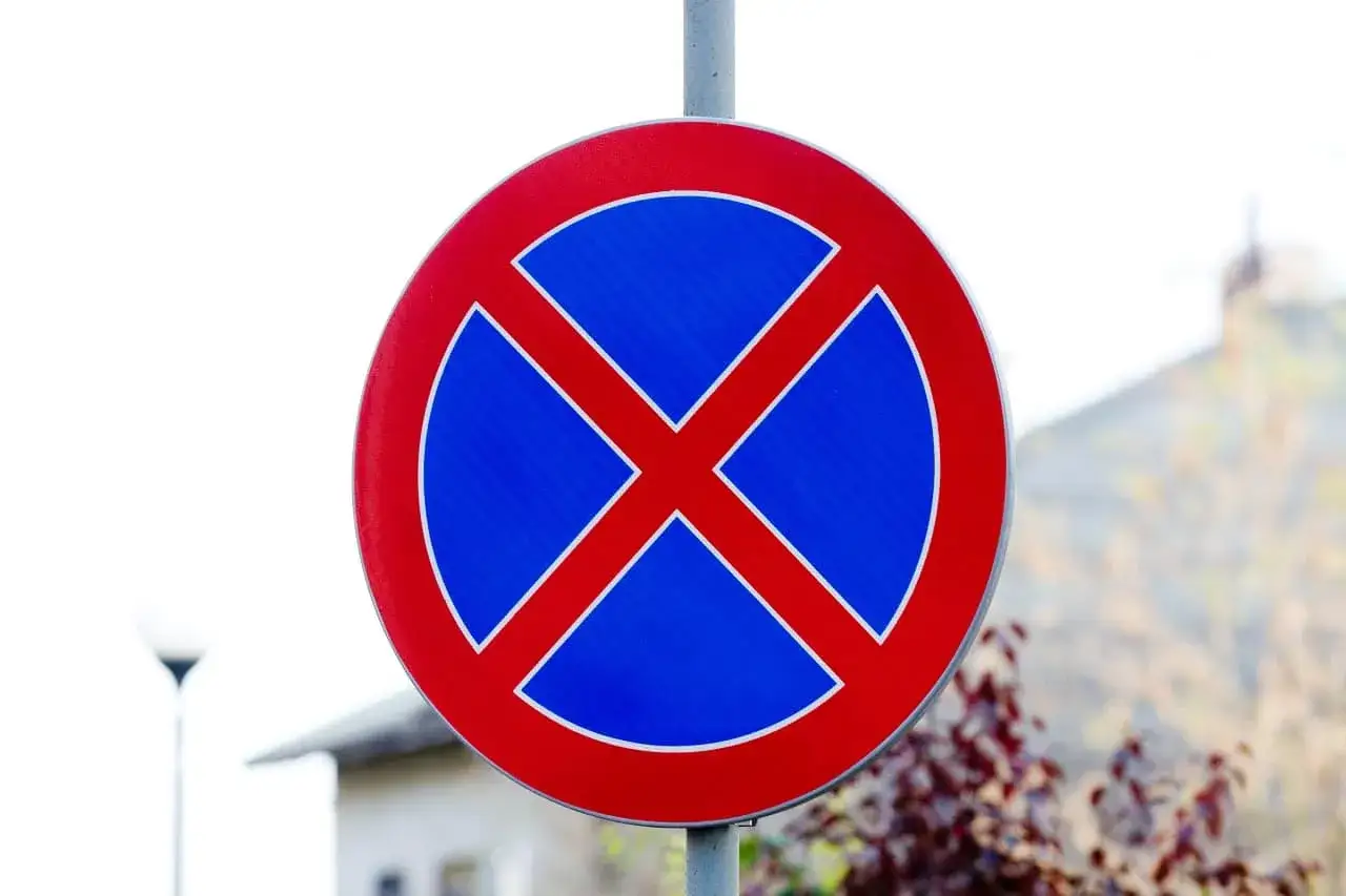 Znak drogowy informujący o **zakazie postoju**. Czerwone X na niebieskim tle.