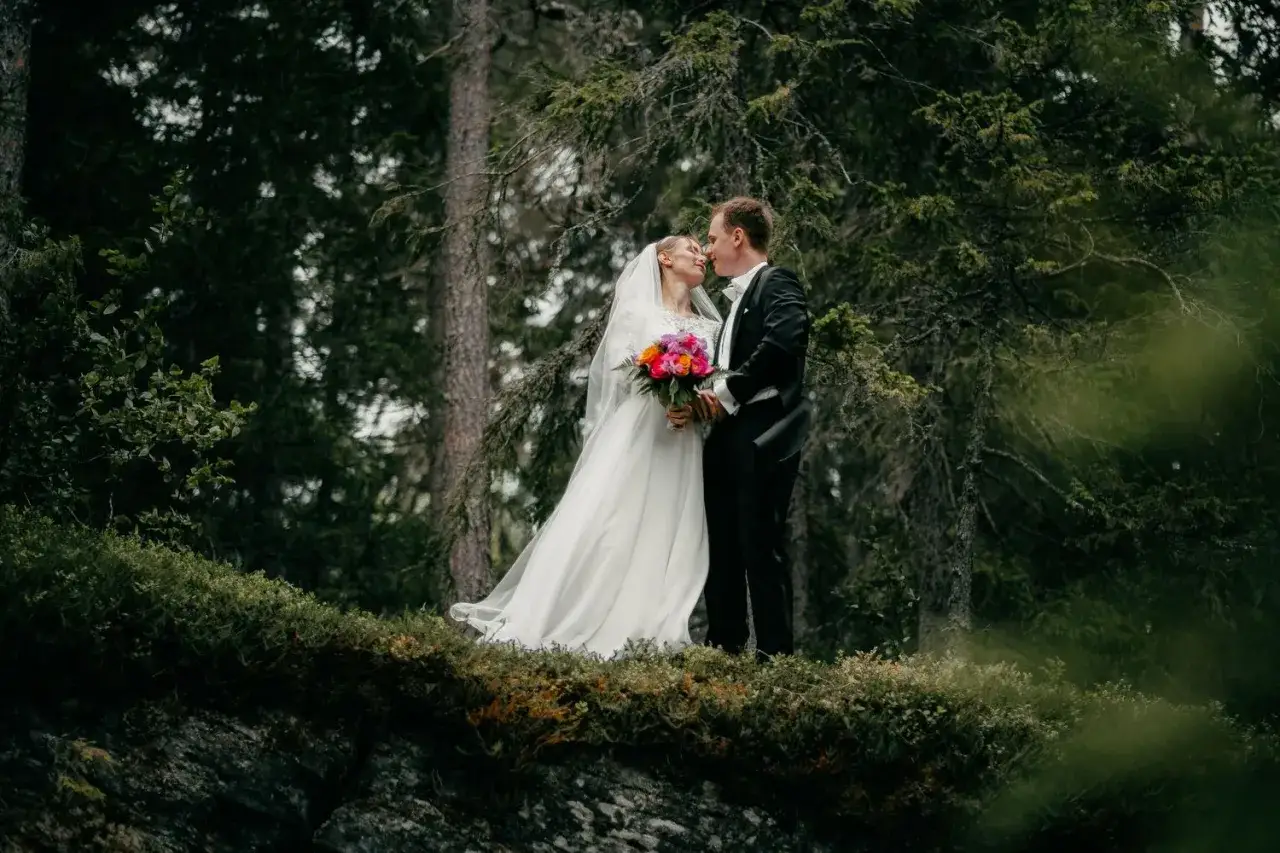 Bröllopspar, Frida Torgeby och hennes man, kysser varandra i en skog. Hon bär en vit klänning och han en svart kostym.
