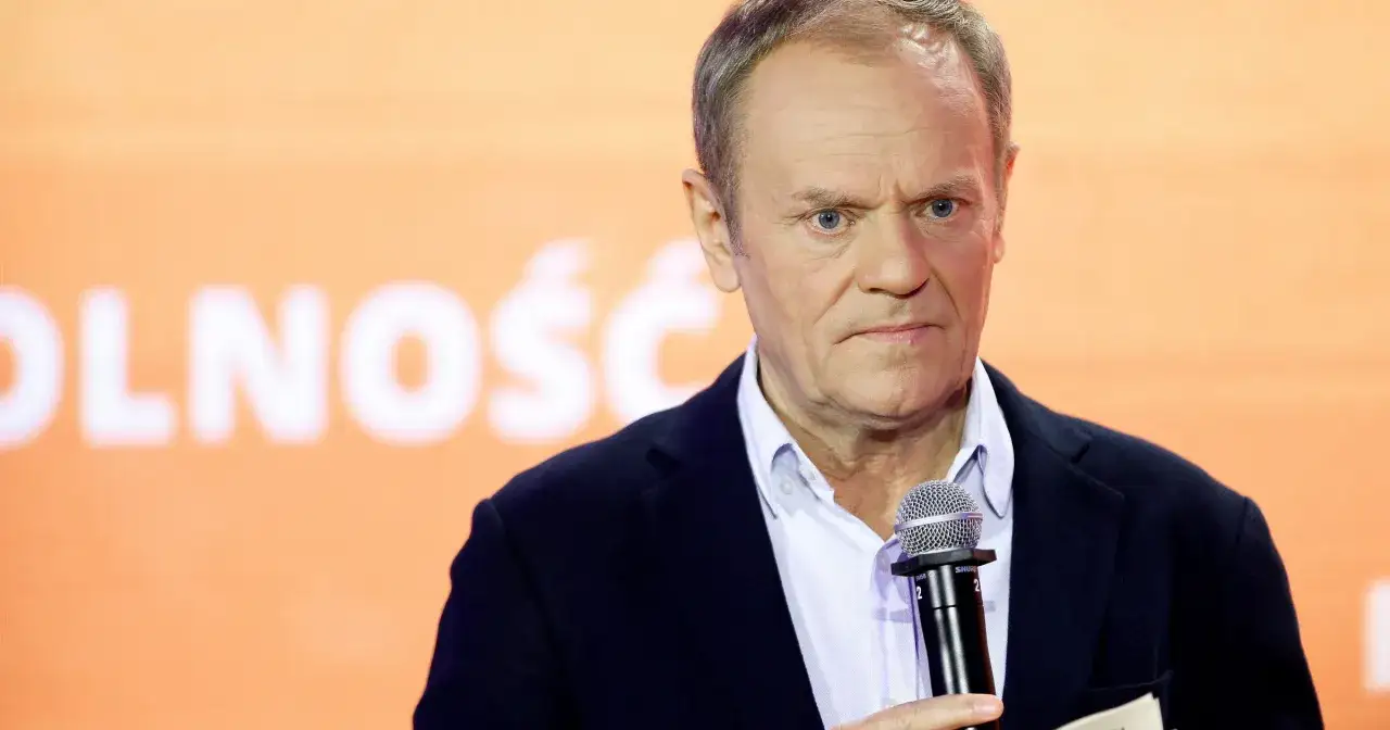 Donald Tusk, polityk o niemieckim pochodzeniu, przemawia do mikrofonu na tle pomarańczowego banera z napisem 