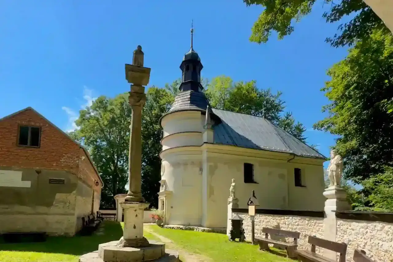 Kamienny obelisk i biała kaplica na terenie pustelni błogosławionej Salomei, otoczone zielenią i błękitnym niebem.