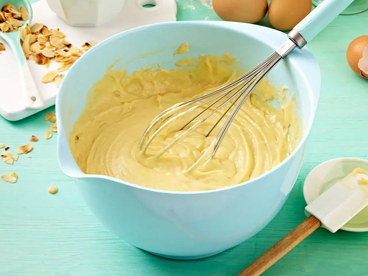 Ein hellblauer Schüssel mit Kuchenteig und Schneebesen. Daneben Mandelsplitter und Eier. Perfekt für ein Kuchenteig Rezept.