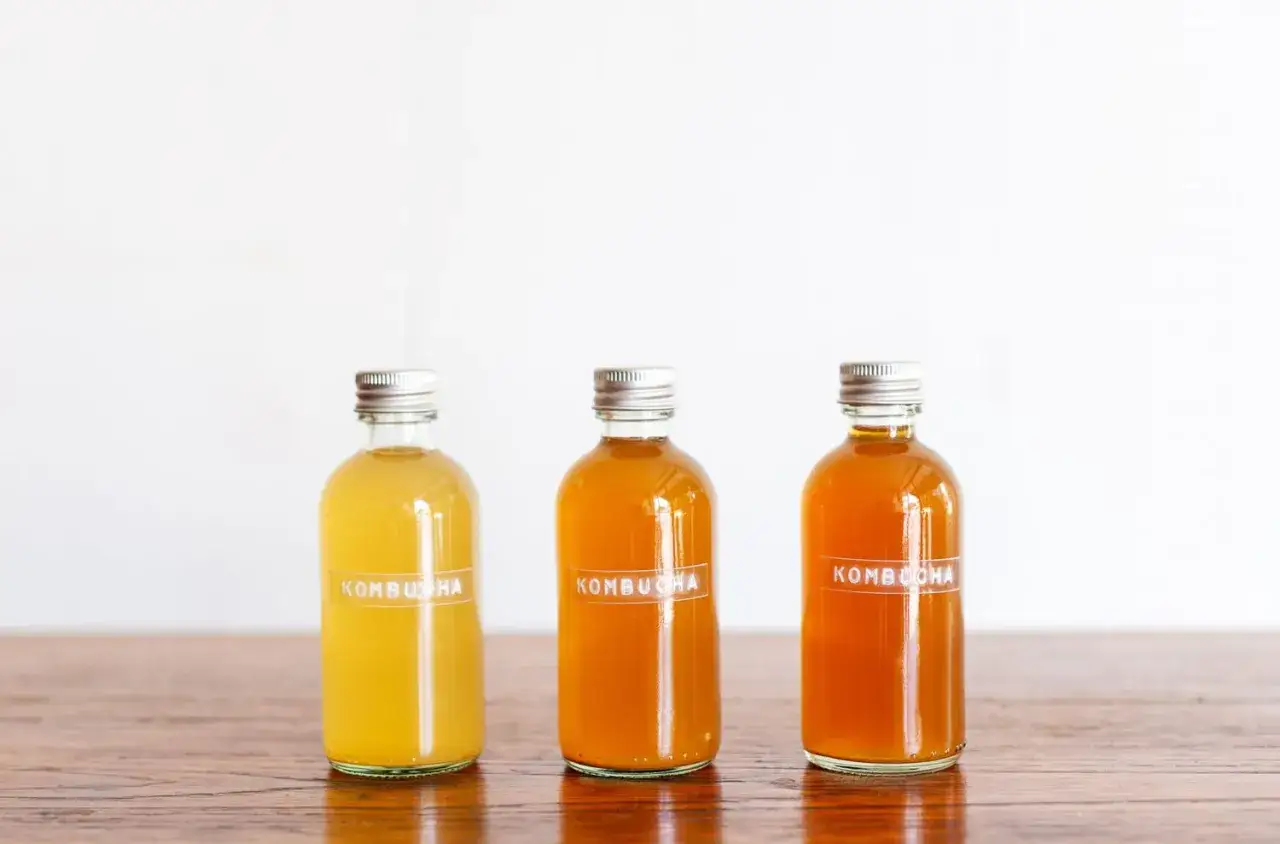 Trois bouteilles de kombucha artisanal aux teintes ambrées et dorées, alignées sur une table en bois.