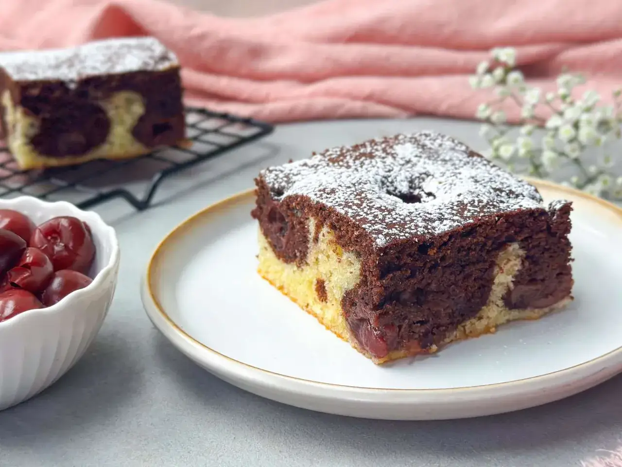 Ein St&uuml;ck saftiger Marmorkuchen mit Kirschen, best&auml;ubt mit Puderzucker. Daneben eine Schale mit frischen Kirschen.