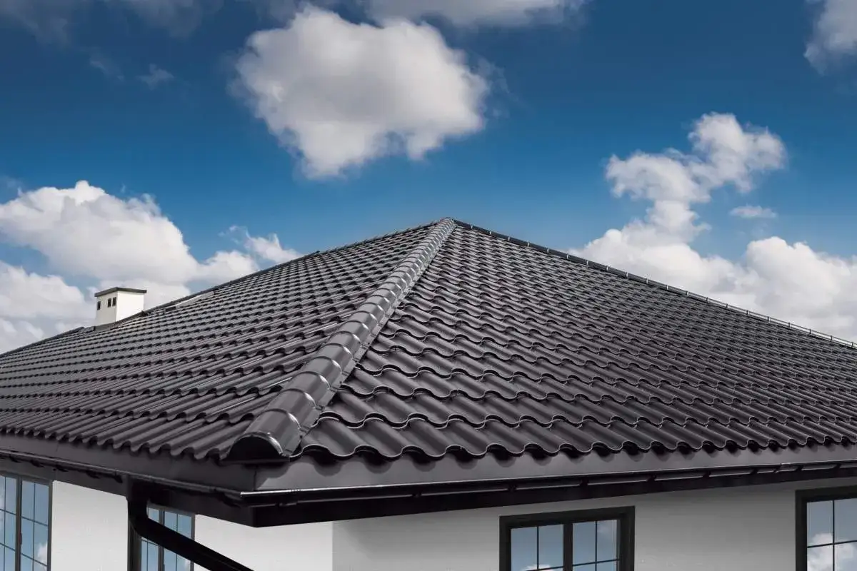 Nowy, czarny dach czterospadowy z dachówki ceramicznej błyszczy w słońcu. Nad nim błękitne niebo z białymi chmurami.