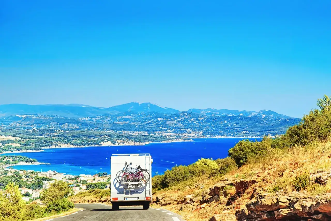 Un camping-car avec deux vélos à l'arrière roule sur une route de campagne, offrant une vue magnifique sur une baie bleue et des montagnes. La consommation de carburant est optimisée pour ce voyage.