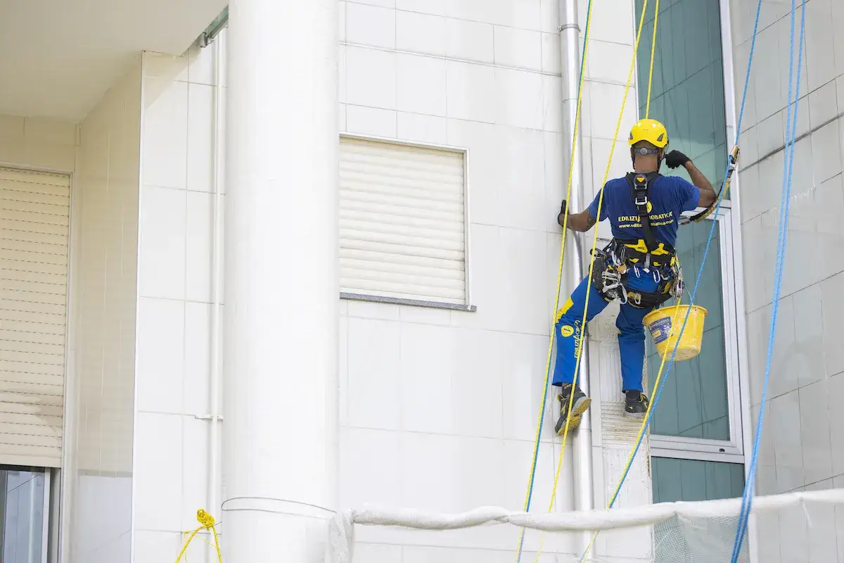 Trabajador en altura, con arn&eacute;s y casco amarillo, desciende por la fachada de un **ite de edificios** blancos, con un cubo amarillo colgando.