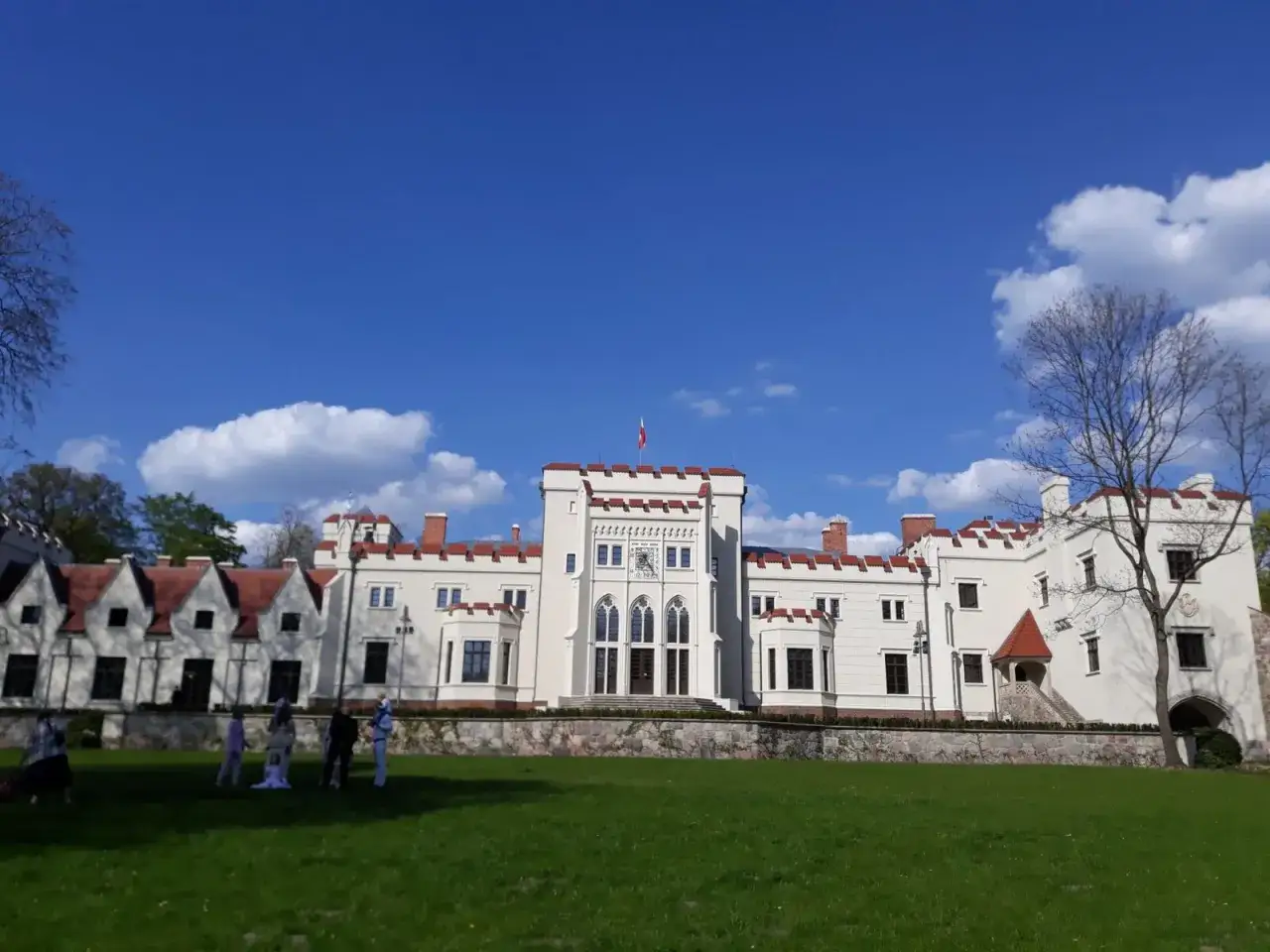 Biały pałac z czerwonym dachem i wieżyczkami, otoczony zielonym trawnikiem. W oddali widać kilka osób spacerujących po parku w Jarocinie.