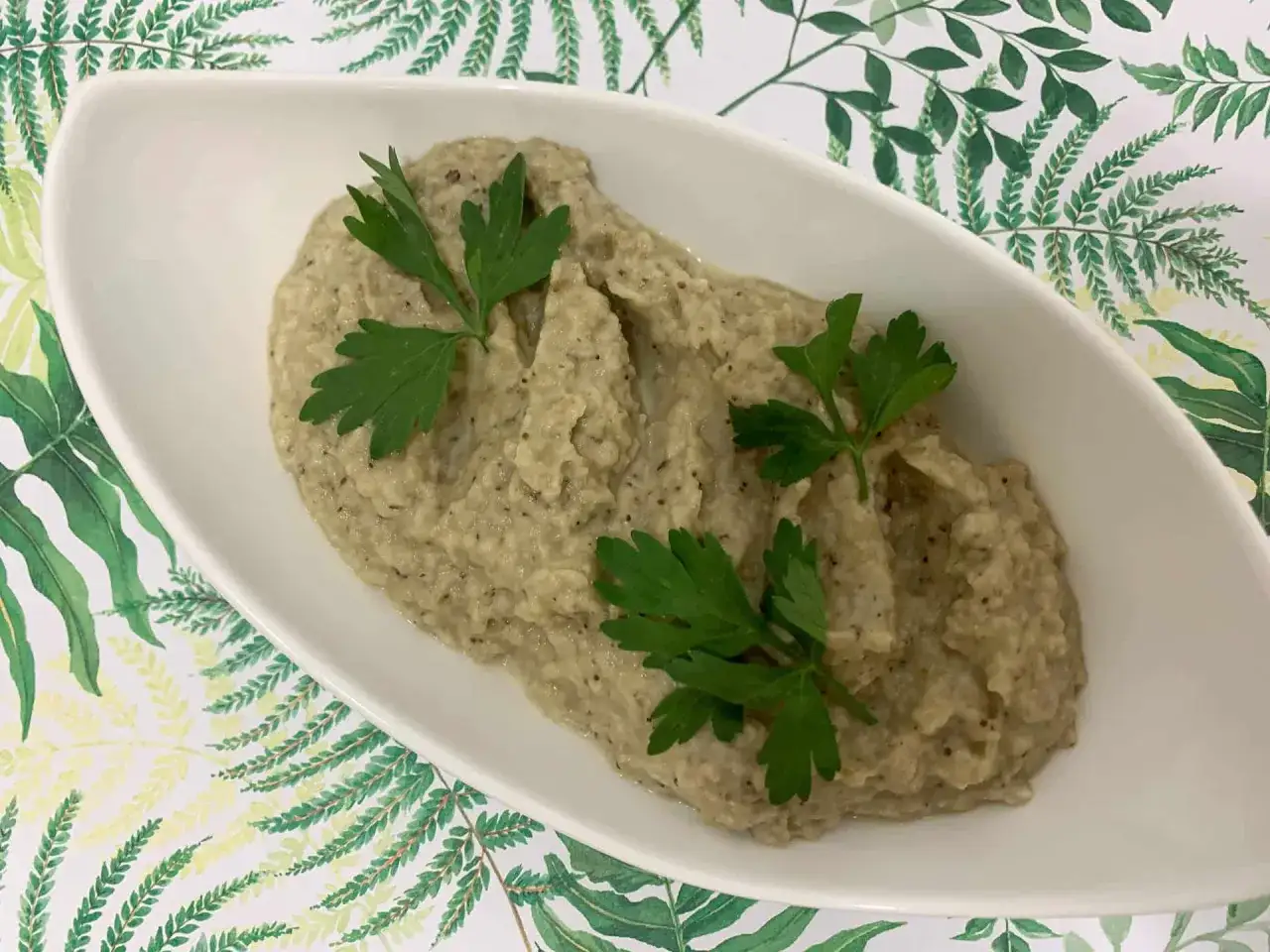Puszysta pasta z bakłażana, udekorowana świeżą natką pietruszki, podana w białej miseczce na tle zielonych liści.
