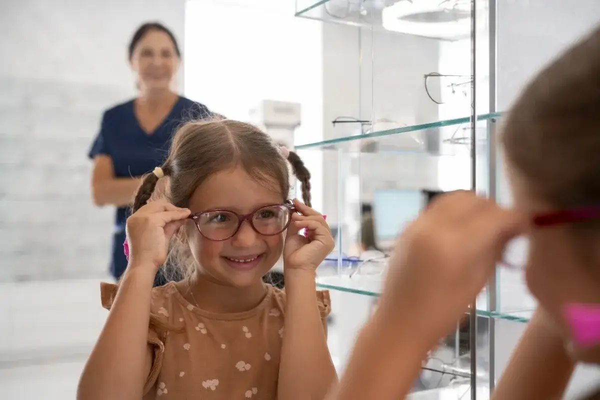 Uśmiechnięta dziewczynka przymierza okulary w salonie optycznym. Zastanawia się, czy można przełożyć szkła do innych oprawek, podczas gdy optometrystka obserwuje ją z tyłu.