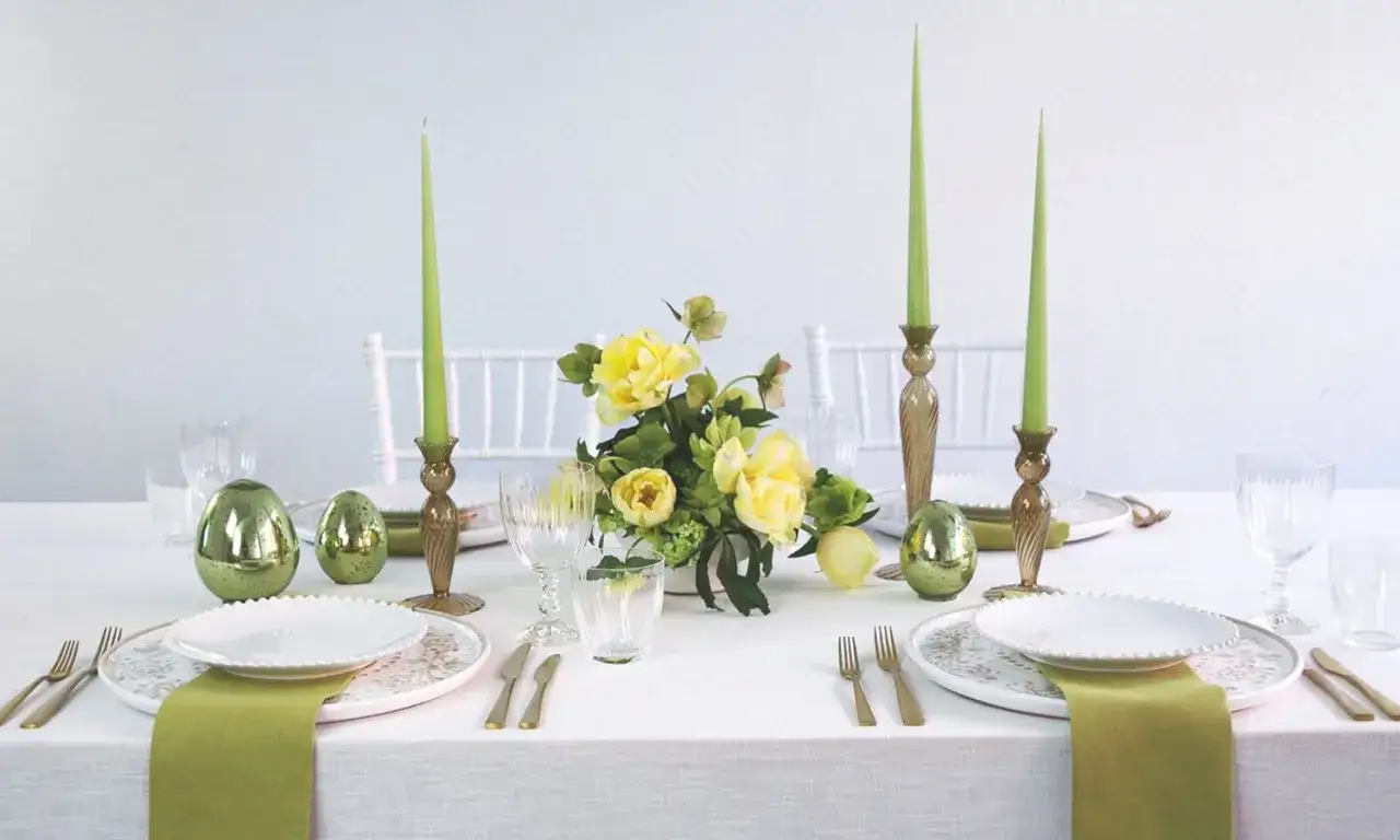 Einfache Tischdeko Ostern: Grüne Kerzen, goldene Eier und gelbe Blumen auf weiß gedecktem Tisch.