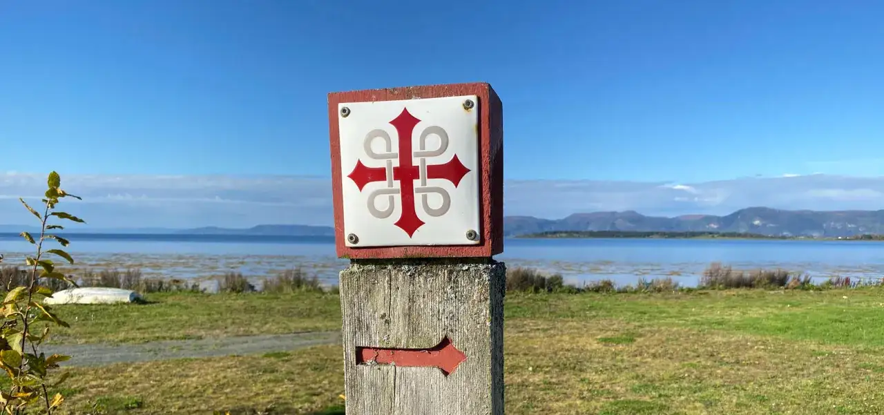 St. Olavsleden-skylt med pil pekar mot en vacker sj&ouml; och berg.