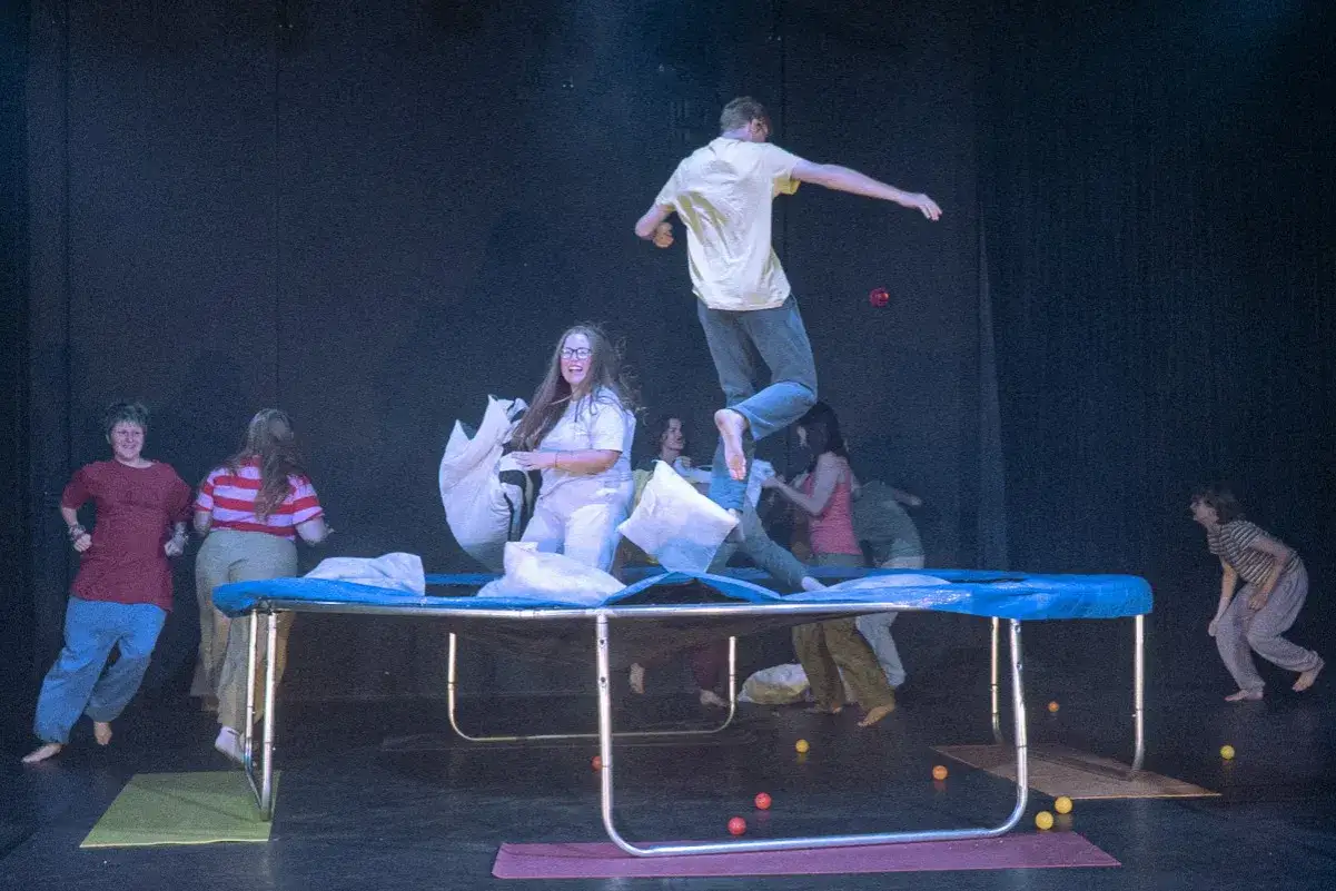 Grupa ludzi skacze na trampolinie, rzucając poduszkami. Scena przypomina tragifarsę, pełną chaosu i radości.