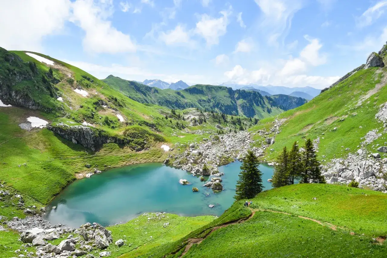 Vue panoramique du lac de Darbon, un joyau turquoise niché au cœur de montagnes verdoyantes, avec des sentiers serpentant sur les pentes et des sommets enneigés à l'horizon.