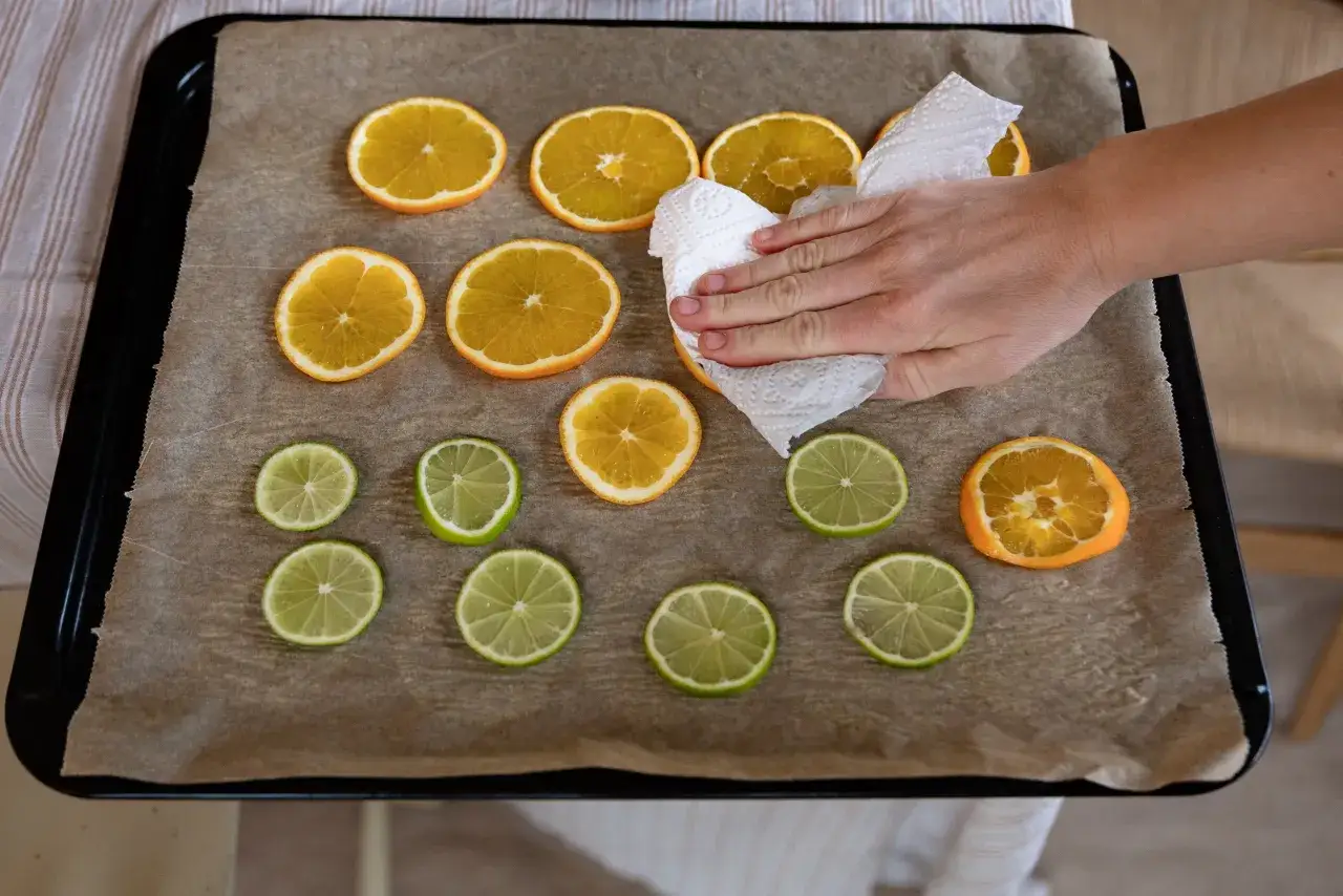 En hand torkar apelsinskivor på en plåt med bakplåtspapper. Citrusfrukter förbereds för att torka.