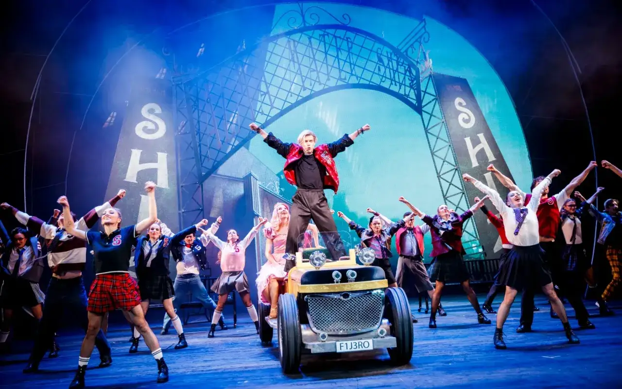 Energetyczny występ na scenie Teatru Roma. Młodzi aktorzy w szkolnych strojach, na pierwszym planie auto z tablicą rejestracyjną F1J3RO. Dyrektor teatru z pewnością dumny z takiego widowiska.