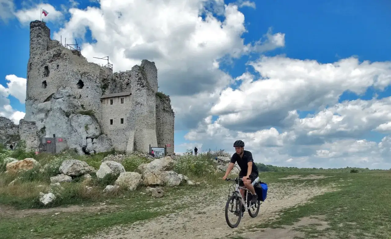 Rowerzysta przemierza malowniczą Jurę Krakowsko-Częstochowską, mijając ruiny zamku na tle błękitnego nieba z chmurami.