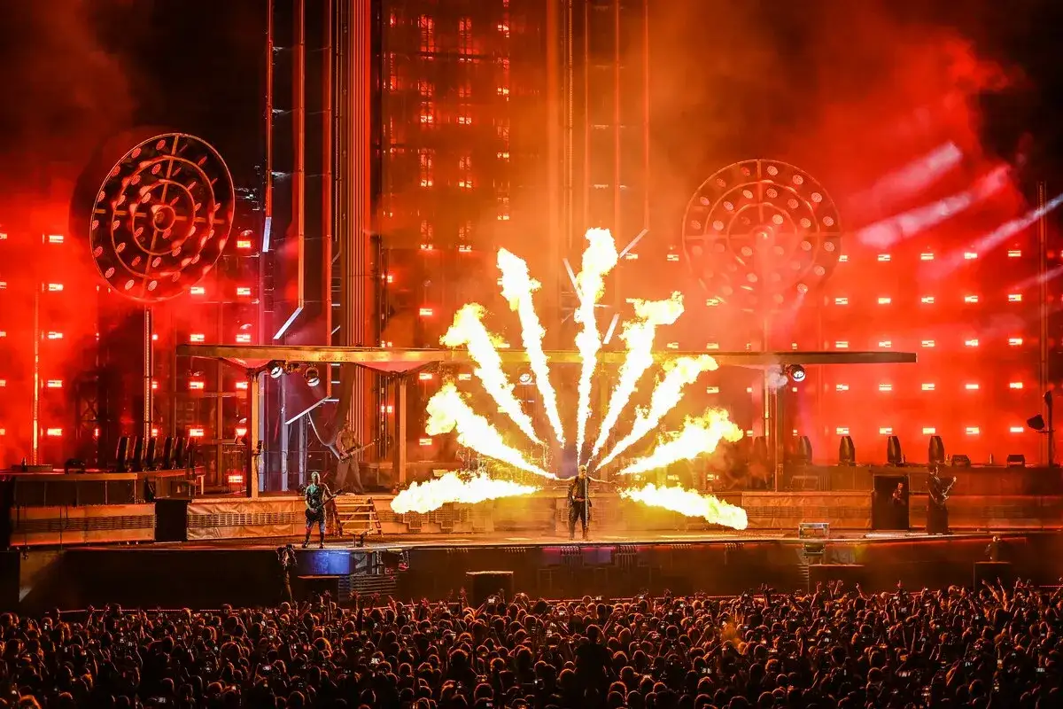 Spectacle pyrotechnique sur scène, un concert Rammstein style de musique, avec des flammes jaillissant en éventail devant une foule immense.