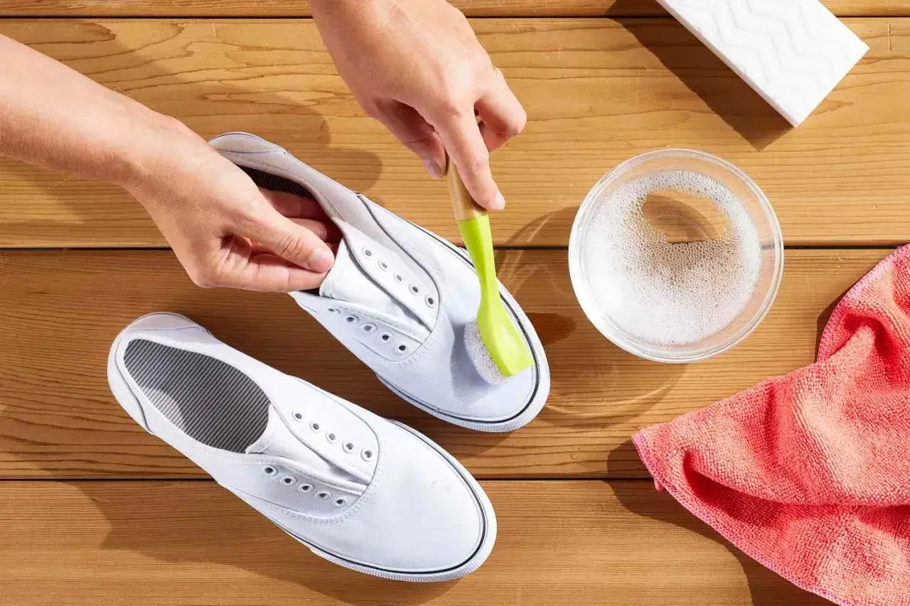 Apprendre comment nettoyer des baskets blanches. Une main brosse une chaussure avec une brosse verte, &agrave; c&ocirc;t&eacute; d'un bol d'eau savonneuse.