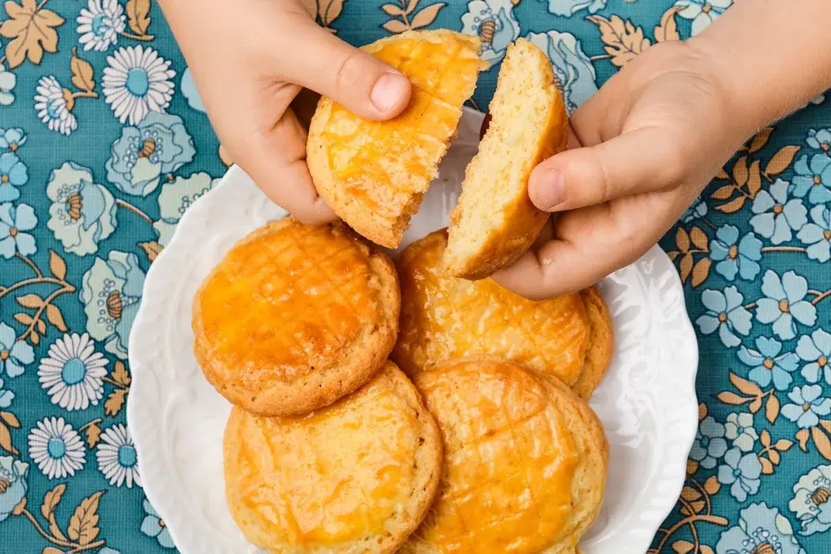 Des mains d'enfant cassent une galette de farine dor&eacute;e. Sur une assiette blanche, d'autres galettes attendent, sur un tissu fleuri bleu.