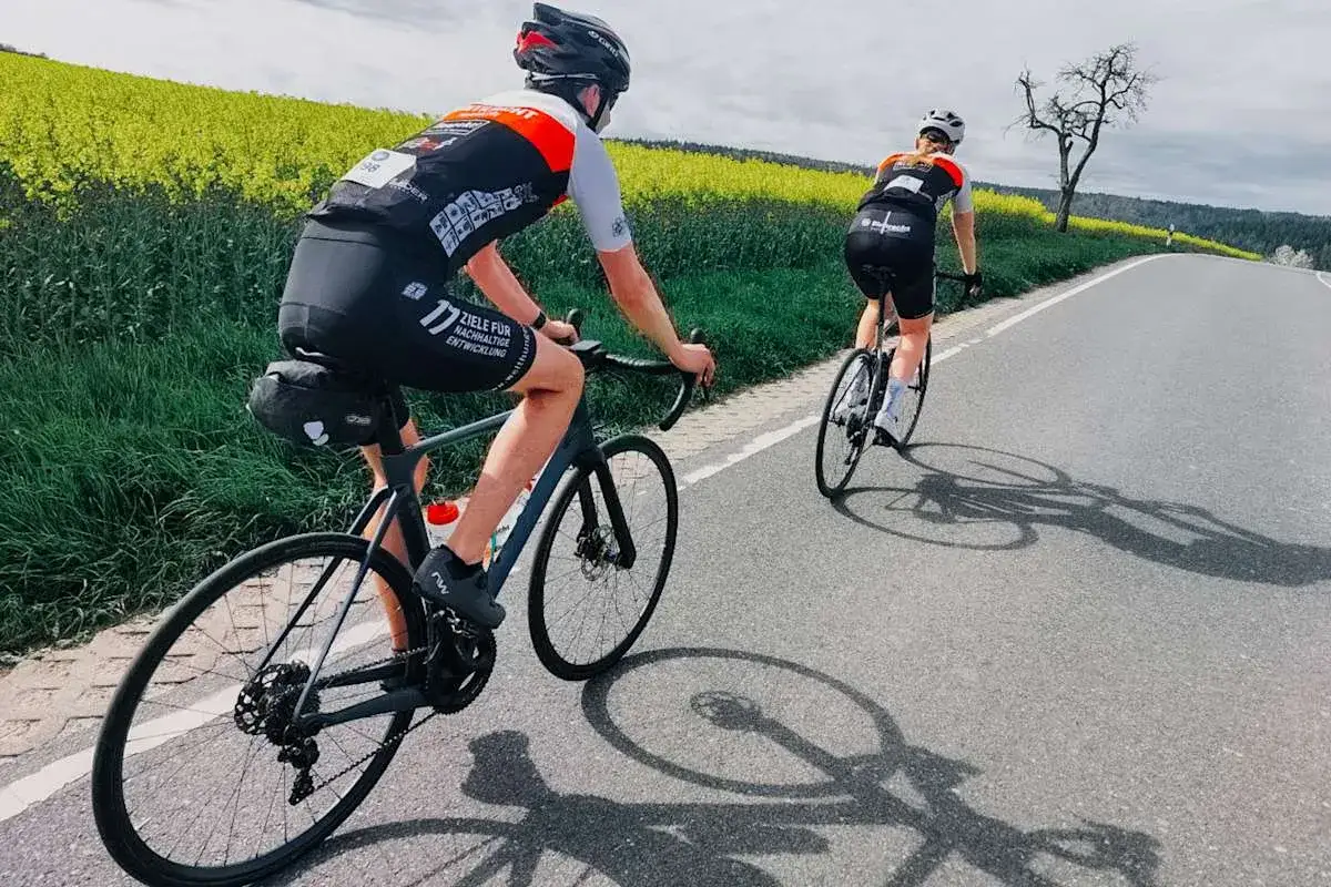 Zwei Radfahrer auf einer Straße, umgeben von Rapsfeldern. Auf dem Bein des vorderen Fahrers steht